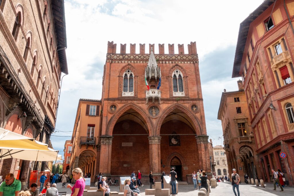A lively street scene in front of a historic palazzo in Italy with diverse people enjoying a sunny day.